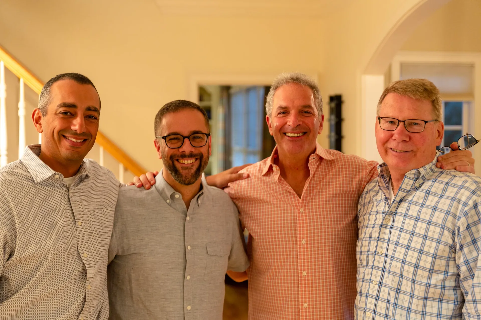 Four smiling men standing shoulder to shoulder with their arms around each other, posing for a group photo