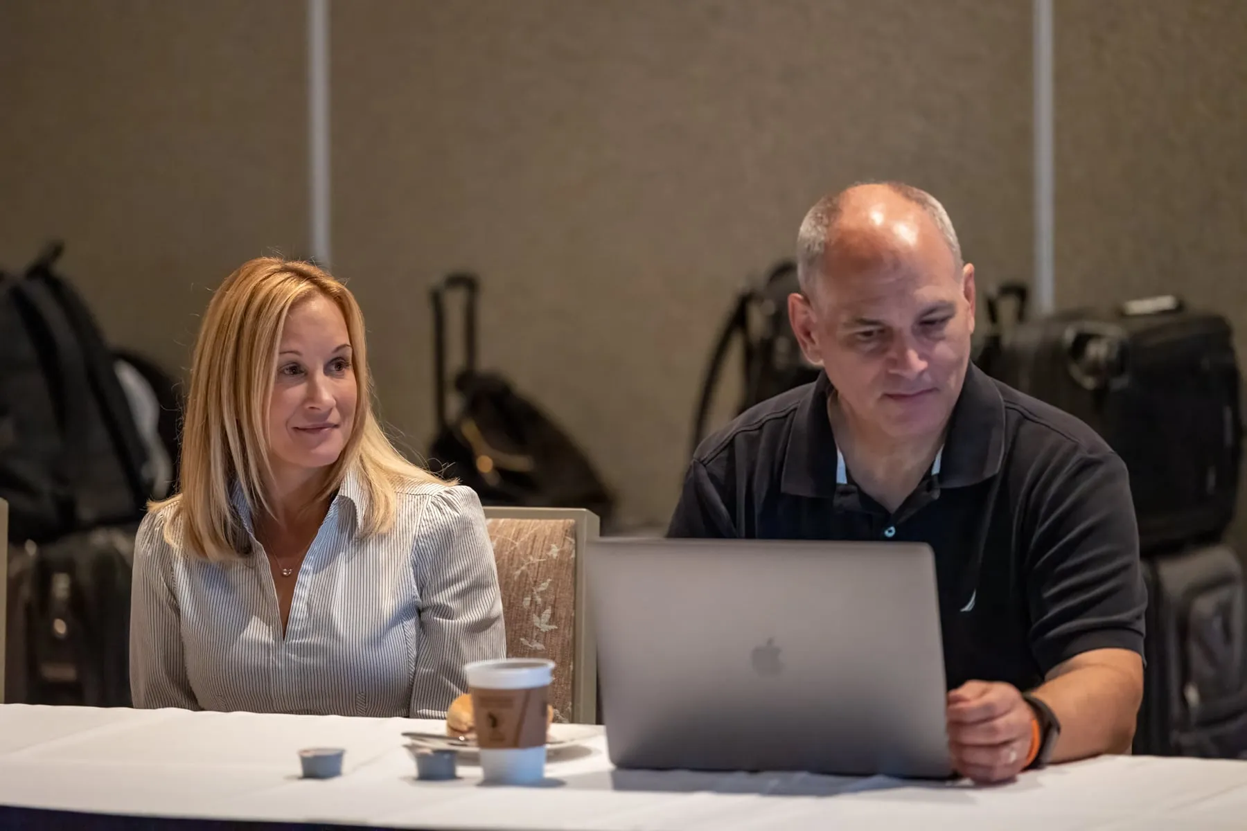 A woman and a man seated at a conference table, he working on a laptop while she looks on