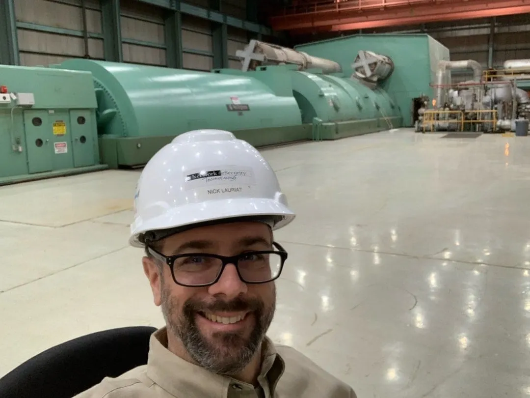 Smiling bearded man with glasses in a white hard hat standing in a power plant turbine hall