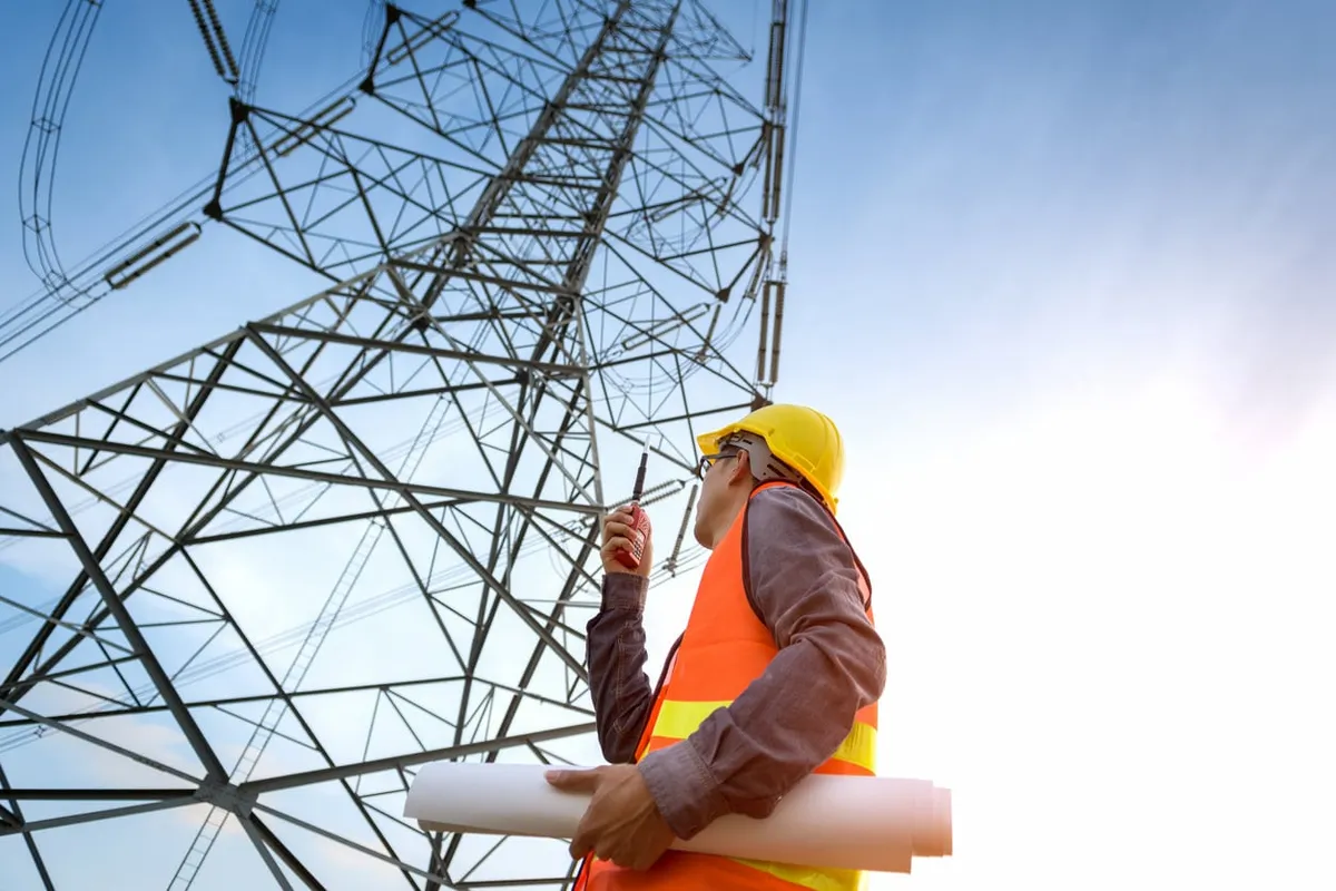 Worker in a hard hat and orange vest holding rolled plans and a radio, looking up at a high-voltage transmission tower