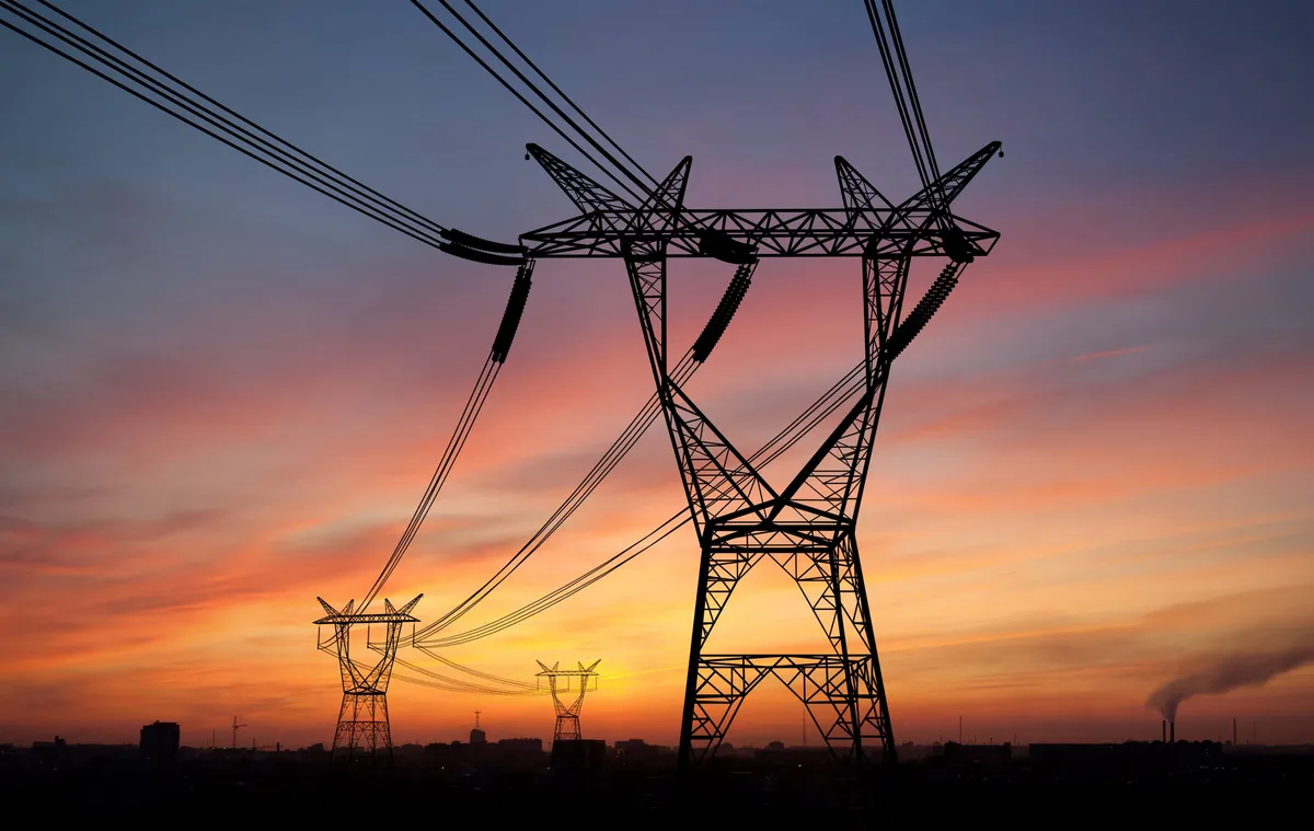 Power line tower and cables silhouetted against an orange sunset over a city skyline