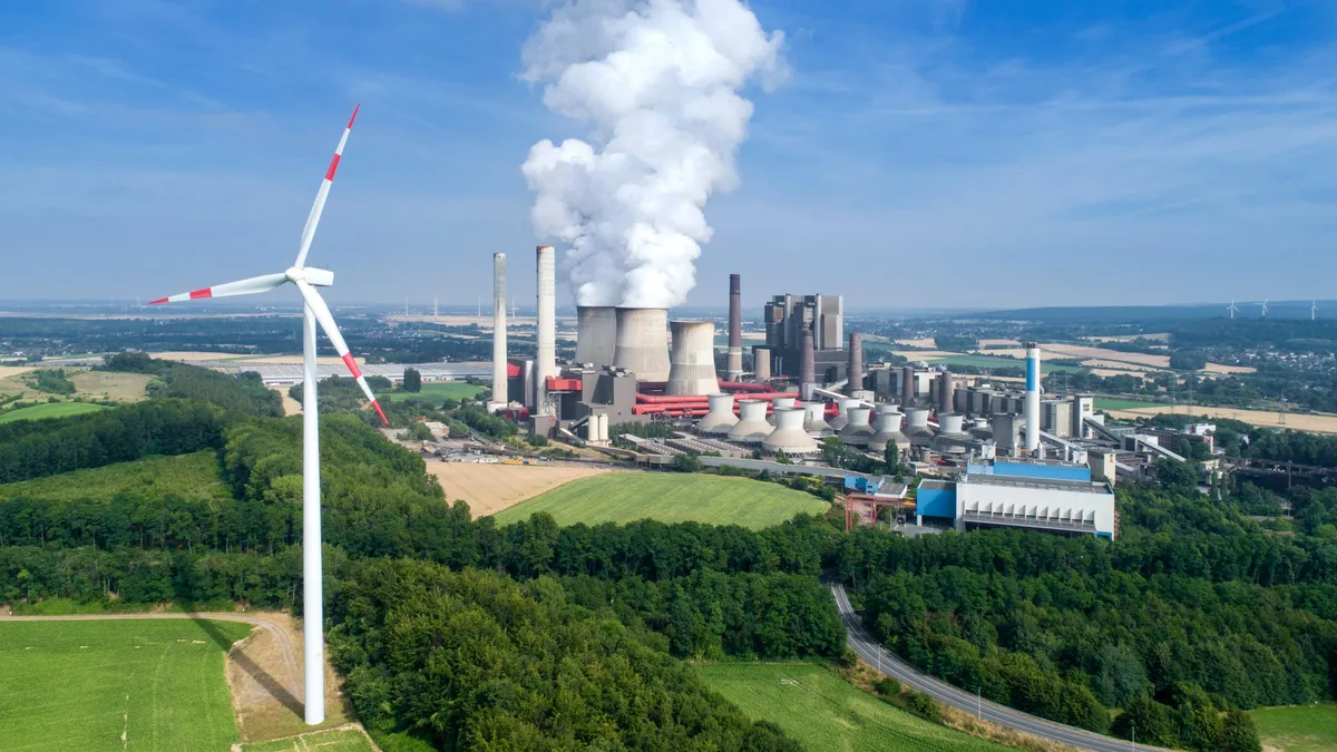 Power plant with steaming cooling towers beyond forest and fields, a wind turbine in the foreground