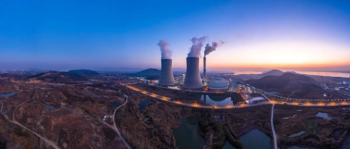 Aerial dusk view of a power station with steaming cooling towers beside a lit coastal road