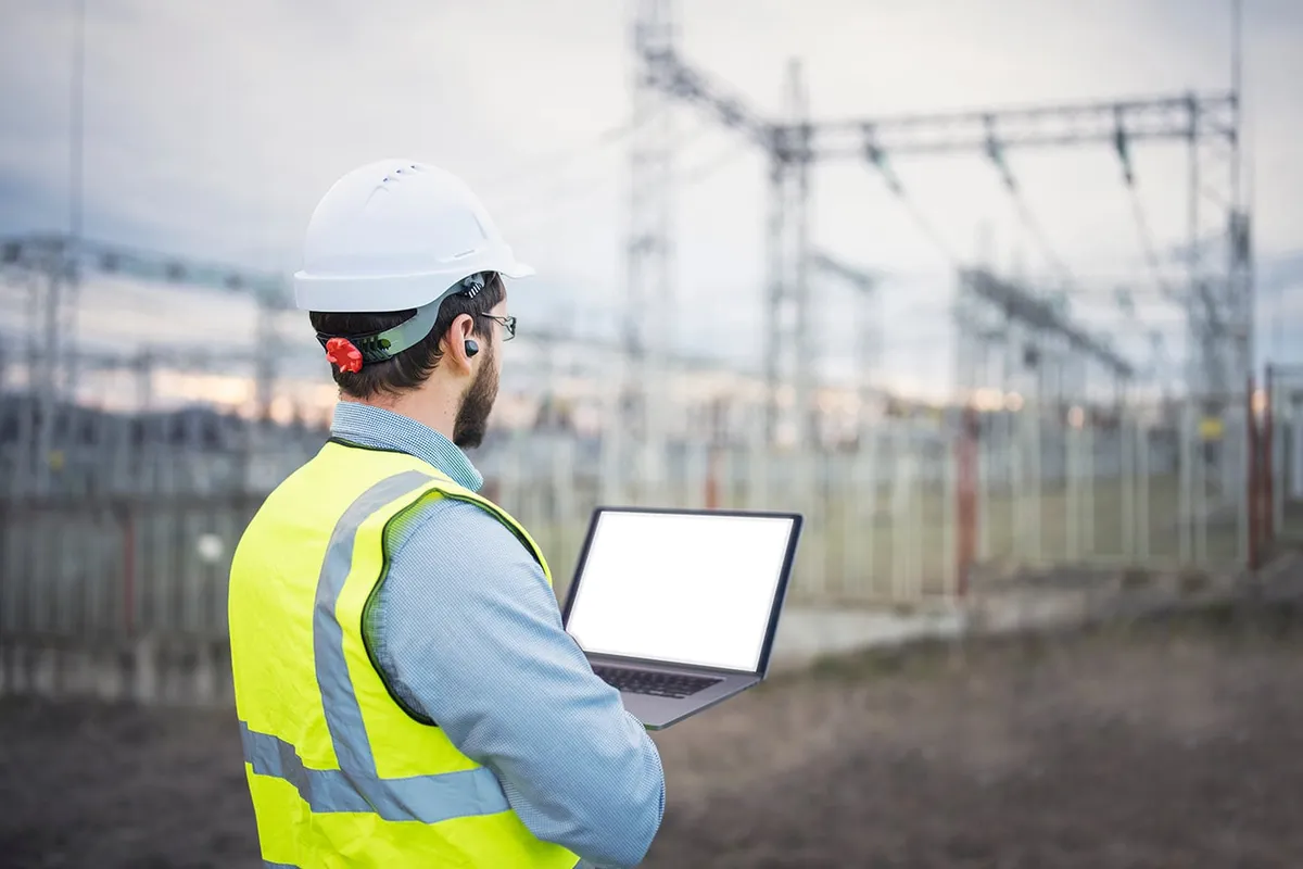 Engineer in a white hard hat and yellow safety vest using a laptop outdoors at an electrical substation