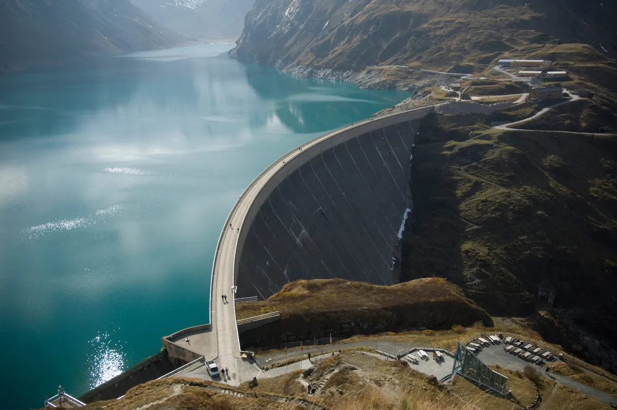 Curved concrete dam holding back a turquoise mountain reservoir, seen from above