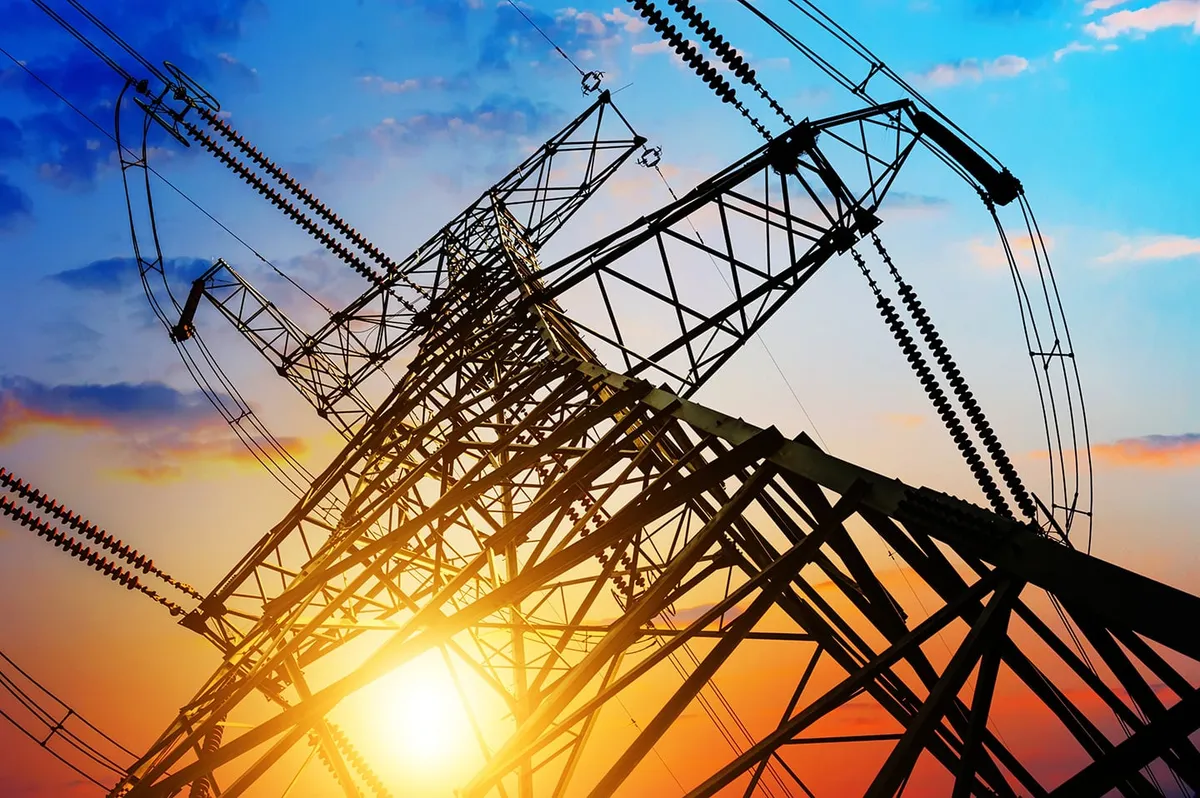 High-voltage transmission tower silhouetted against a bright setting sun and blue sky
