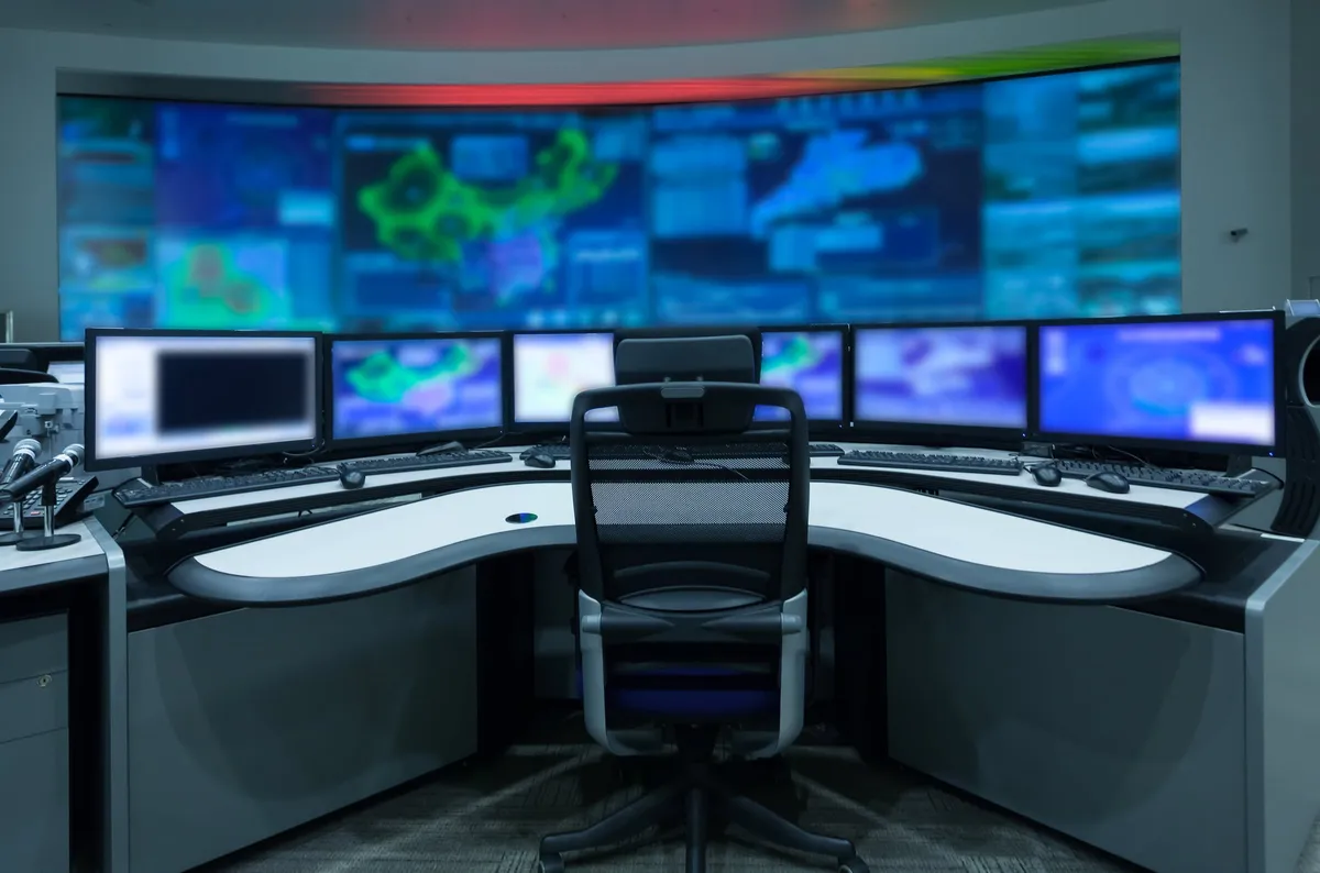 Utility control room with a wall of monitoring screens above empty operator workstations