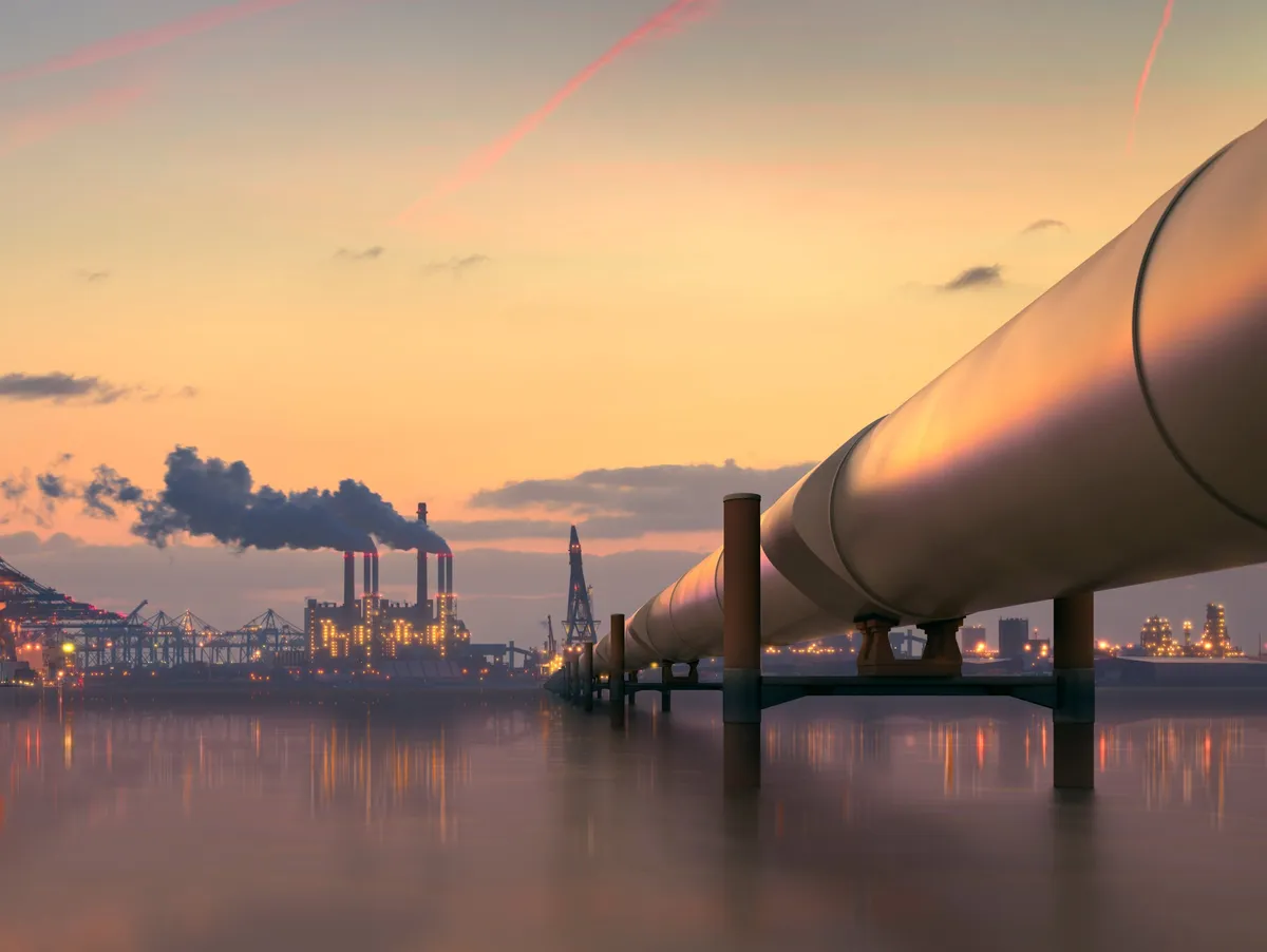 Large pipeline crossing water toward an industrial plant silhouetted at dusk