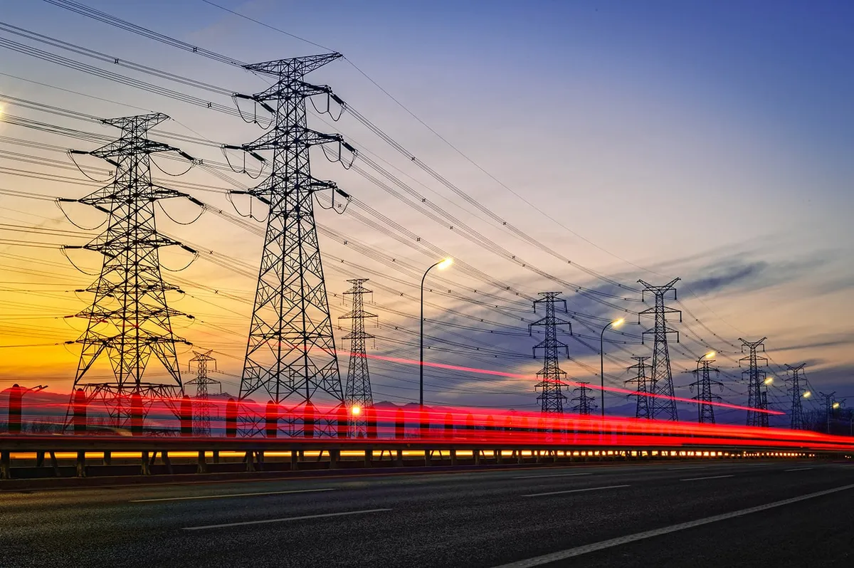 Rows of high-voltage towers beside a road at sunset, with red light trails from passing traffic