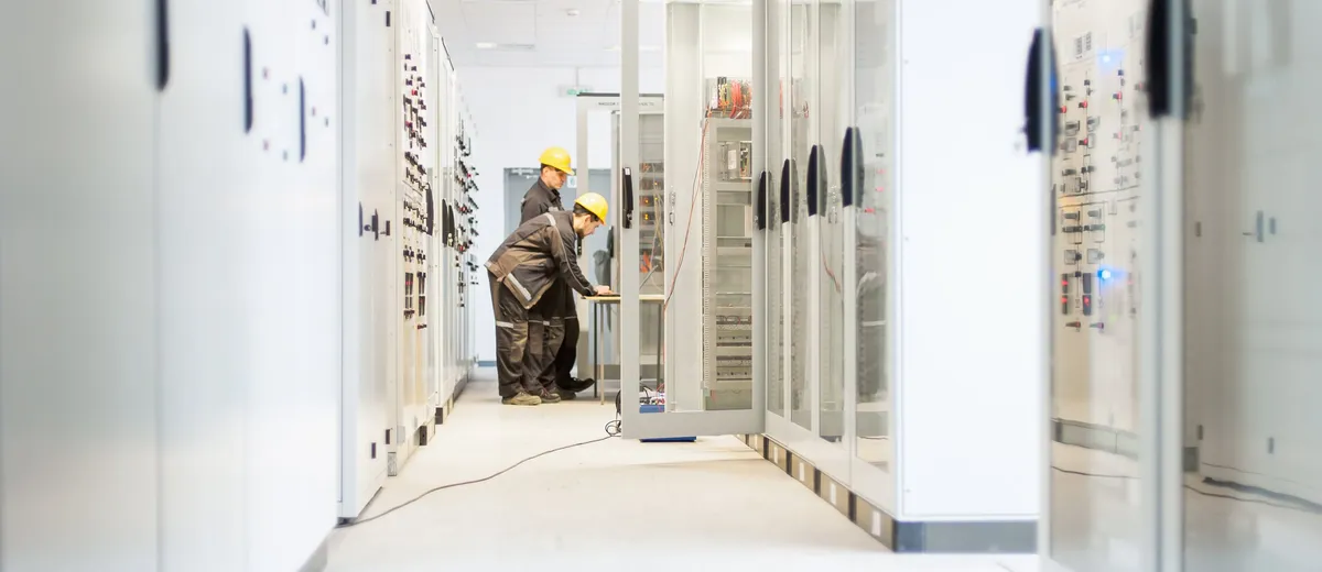 Two workers in hard hats at an open electrical cabinet in a bright equipment room