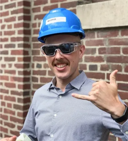 Smiling man in a blue hard hat and safety glasses making a shaka hand sign in front of a brick wall