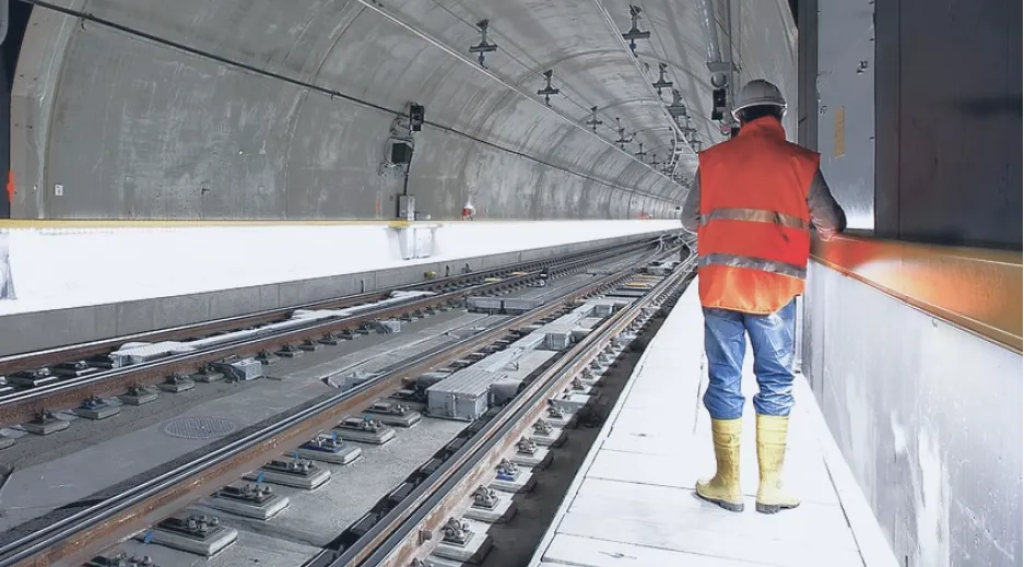 Worker in an orange vest and hard hat walking beside railway tracks inside a tunnel